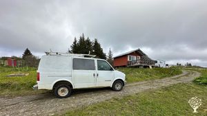 Foreground is white van. Background is outhouse to left and red cabin packhouse to the right.